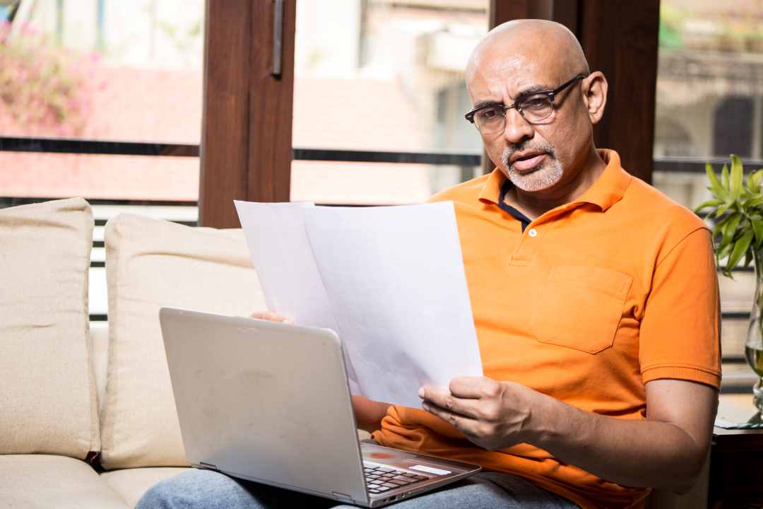Middle aged man reading papers with laptop on couch