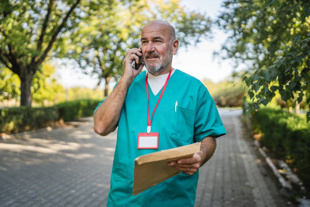 Male doctor wearing scrubs on the phone and holding a document outdoors  